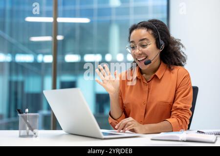 Une jeune femme d'affaires passe un appel vidéo à son bureau, en utilisant un ordinateur portable et portant un casque. Elle apparaît amicale et professionnelle, incarnant le travail à distance et la communication. Banque D'Images