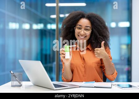 Jeune femme d'affaires montrant le plaisir d'utiliser un spray de gorge à son lieu de travail. Elle sourit avec confiance, donne le pouce levé, et est assise à un bureau avec un ordinateur portable et un téléphone Banque D'Images