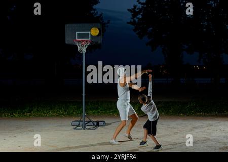 Un père joue au basket-ball avec son jeune fils la nuit dans un parc éclairé par une lumière vive Banque D'Images