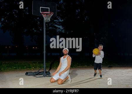 Un père joue au basket-ball avec son jeune fils la nuit dans un parc éclairé par une lumière vive Banque D'Images