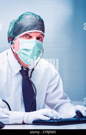 Médecin portant un masque chirurgical, une résille à cheveux et des gants, tapant sur un clavier tout en établissant un contact visuel avec la caméra, mettant en vedette un stéthoscope autour de hi Banque D'Images