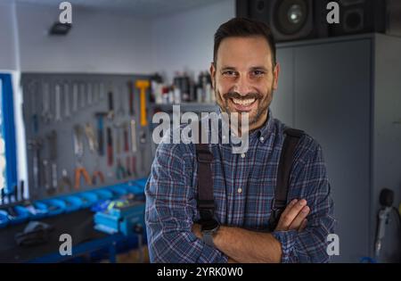 Portrait de beau réparateur portant debout avec les bras croisés dans l'atelier, outils accrochés au mur en arrière-plan Banque D'Images