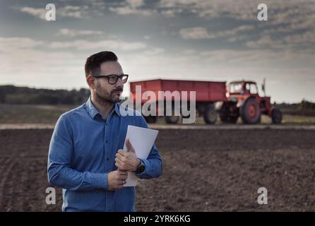 Portrait d'homme d'affaires confiant tenant le cahier devant le tracteur avec remorque dans le champ en automne. Agent bancaire et concept de prêt pour petite entreprise Banque D'Images