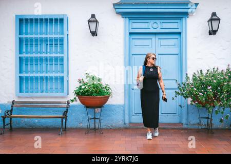 Une femme à la mode dans une robe noire et des baskets passe devant une porte bleue de style colonial et une fenêtre ornée de fleurs. La scène vibrante mélange Trad Banque D'Images
