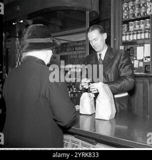 Dans la boutique en 1942. Assistant masculin dans l'épicerie fine d'Arvid Nordquist sur Birgerjarlsgatan à Stockholm. Nous sommes en 1942 et la seconde Guerre mondiale fait rage en Europe. Malgré la guerre, l'entreprise est en mesure de proposer des épiceries et des délices. La boutique n'est pas encore entièrement passée en libre-service et prend la commande au comptoir. On le voit faire des paquets rangés de l'épicerie. 1942. Kristoffersson ref A123-5 Banque D'Images