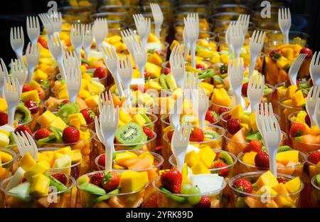 Salade de fruits frais (kiwi, mangue, fraise, etc.) disposée dans des gobelets en plastique avec fourchettes sur un étal de marché. Foyer sélectionné. Concept de restauration rapide sain. Banque D'Images