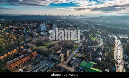 Vue aérienne d'un quartier résidentiel avec des immeubles de grande hauteur, des maisons mitoyennes et une ligne d'horizon lointaine de la ville sous un ciel partiellement nuageux. Une chaussée avec movi Banque D'Images