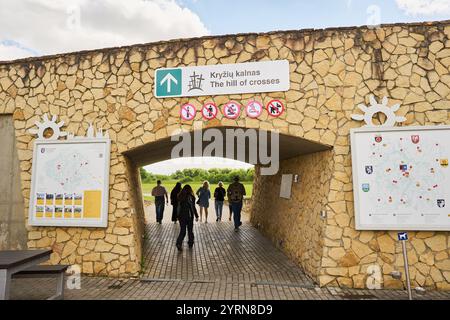 Les visiteurs traversent une arche en pierre menant à la colline des croix, en Lituanie, marquée par des panneaux informatifs et des cartes dans un paysage pittoresque. Banque D'Images