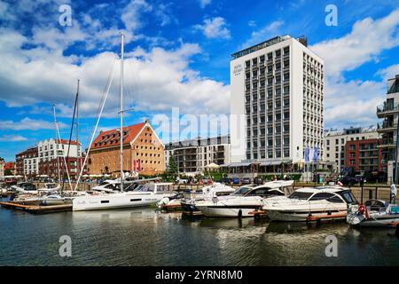 Une scène de marina dynamique à Gdansk avec divers bateaux amarrés à côté de bâtiments modernes et historiques sous un ciel bleu avec des nuages moelleux. Banque D'Images