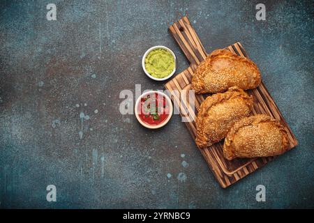 Délicieuses empanadas cuites au four avec graines de sésame servies sur planche de bois avec sauces, pâtisserie traditionnelle espagnole salée Banque D'Images