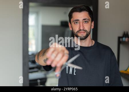 Beau gars de salon de coiffure en T-shirt noir tient des ciseaux et regarde la caméra. Banque D'Images