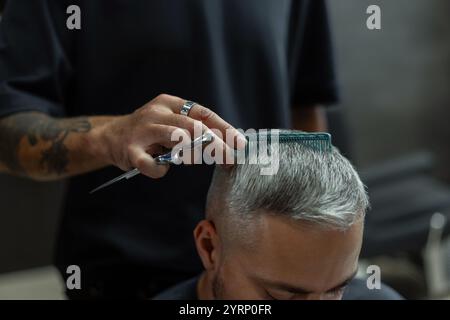 Cool Barber Man coupe un homme aux cheveux gris dans un studio de coiffure. Main masculine avec tatouage tient des ciseaux et un peigne dans le salon de coiffure Banque D'Images