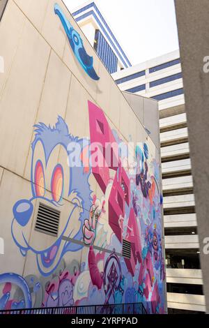 DAS Foto zeigt eine detailreiche und farbenfrohe Street-Art-Komposition an einer Betonwand à Charlotte, Caroline du Nord. Zu sehen ist ein überdimensionaler Cartoon-Charakter mit großen, bunten Augen, der durch abstrakte und surrealistische Elemente ergänzt wird. Dominierende Farben sind Blau, Pink und Lila, die die die Wand mit einer lebhaften Energie erfüllen. In der Mitte des Kunstwerks ragt das Wort PEACE in großen, kantigen Buchstaben hervor, begleitet von verspielten Symbolen und Charakteren. Der obere Bereich zeigt einen stilisierten blauen Vogel, der das Werk mit einem Hauch von Natur berei Banque D'Images