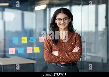 Une jeune femme d'affaires confiante se tient les bras croisés dans un environnement de bureau moderne. Elle porte des lunettes et des sourires, incarnant le professionnalisme et le leadership. Banque D'Images