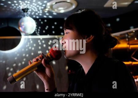 Vue latérale d'une jeune femme asiatique tenant un micro, chantant karaoké dans KTV Banque D'Images