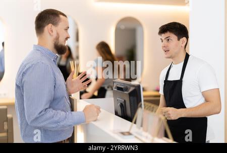 Styliste masculin se tient près du bureau de réception du salon de coiffure, communique avec le visiteur masculin Banque D'Images