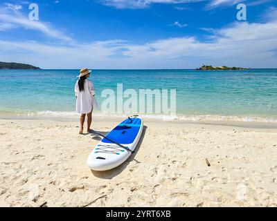 Profitant d'une journée tranquille sur le sable doux de l'île de Samae San, un paddleboard repose à côté d'un aventurier solitaire contemplant les eaux cristallines. Une évasion parfaite dans la nature vous attend sous le ciel bleu. Banque D'Images