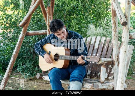 Jeune musicien strumming guitare sur balançoire rustique dans un jardin luxuriant Banque D'Images