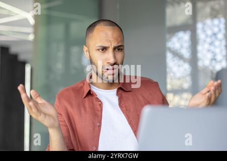 Un employé de bureau perplexe est assis à son bureau, faisant des gestes dans la confusion tout en regardant son écran d'ordinateur. La scène suggère une réunion virtuelle problématique ou un problème de document inattendu. Banque D'Images