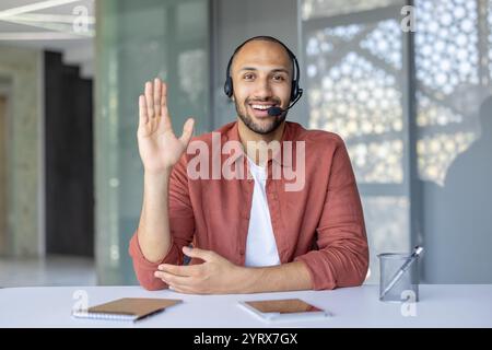 Un homme d'affaires confiant portant un micro-casque participe à une réunion en ligne. Assis à un bureau avec un bloc-notes et un téléphone, il communique efficacement. Idéal pour les concepts d'entreprise et de communication. Banque D'Images