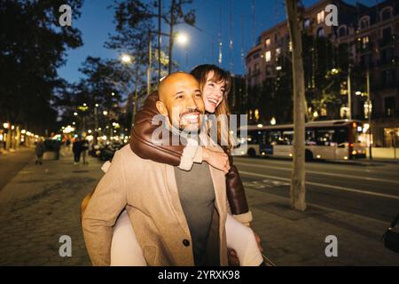 Un couple souriant se régalera dans une balade en piggyback le long d'une rue animée de la ville la nuit, améliorant l'ambiance romantique de la soirée. Banque D'Images