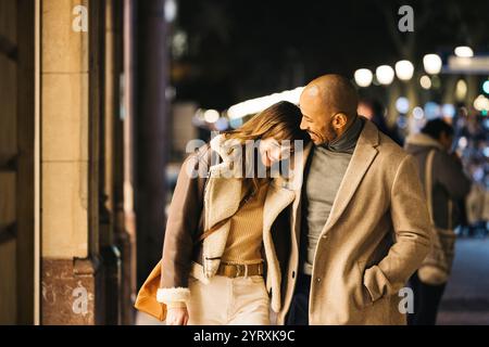 Un couple aimant partage un moment joyeux tout en se promenant dans une rue de la ville la nuit. Ils profitent d'une excursion shopping en souriant et en embrassant l'intima Banque D'Images