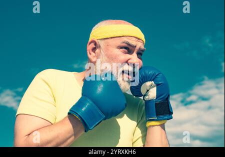 Hommes seniors combattant des poses. Grand-père faisant un entraînement de boxe le matin. Portrait d'un boxeur senior déterminé sur fond de ciel bleu. Banque D'Images