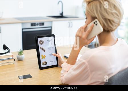 Femme caucasienne commandant de la nourriture en ligne en utilisant la tablette tout en parlant au téléphone. Elle parcourt le site Web de livraison de nourriture dans la cuisine moderne, en se concentrant sur la commodité et la technologie. Banque D'Images
