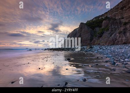 Les vagues se jettent doucement sur le rivage sablonneux tandis que le soleil se couche derrière des falaises escarpées, projetant de belles teintes sur le paysage côtier serein de Dana point. Banque D'Images