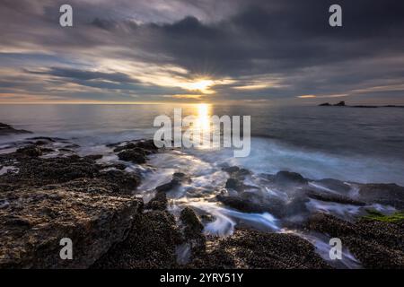 Des vagues douces se battent contre des rivages rocheux tandis qu'un coucher de soleil vibrant projette une lueur chaude sur les eaux tranquilles de Crescent Bay à Laguna Beach, en Californie. Banque D'Images