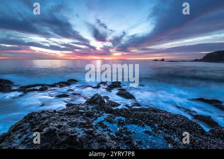 Les vagues s'écrasent doucement contre des rochers accidentés tandis que le ciel se transforme en teintes vibrantes au coucher du soleil à Crescent Bay Beach, Laguna Beach. Banque D'Images