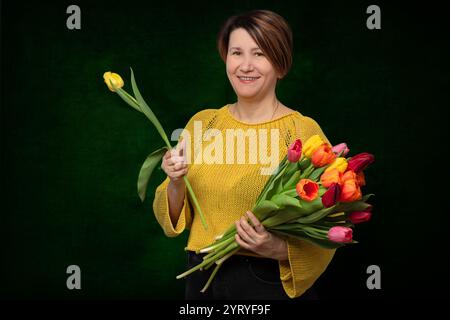 Une belle femme d'âge moyen tient un bouquet de fleurs de tulipes, sourit et regarde la caméra sur un fond sombre Banque D'Images