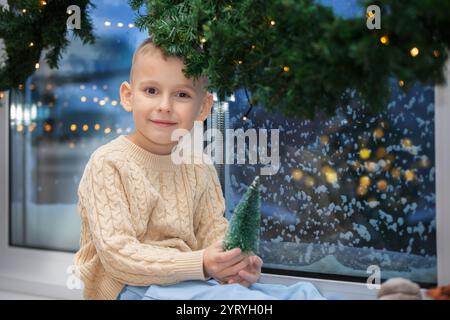 Petit garçon heureux est assis sur le rebord de la fenêtre d'une fenêtre couverte de neige avec décoration de sapin de Noël. Enfant dans la chambre de Noël. Banque D'Images