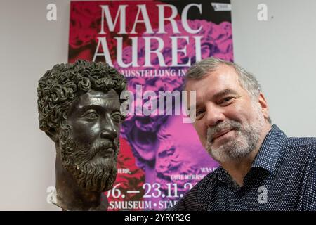 Trèves, Allemagne. 04th Dec, 2024. Marcus Reuter, directeur du Rheinisches Landesmuseum de Trèves, regarde dans la caméra devant une affiche pour l'exposition d'État à côté d'une réplique de la tête de l'empereur romain Marc Aurèle. L’exposition au Rheinisches Landesmuseum et au Stadtmuseum Trèves se déroule du 15 juin au 23 novembre 2025. Crédit : Harald Tittel/dpa/Alamy Live News Banque D'Images