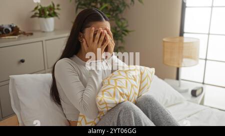 Jeune femme assise dans le lit avec les mains couvrant le visage, regardant en détresse dans un cadre de chambre confortable avec une plante sur une commode et une fenêtre fournissant nat Banque D'Images