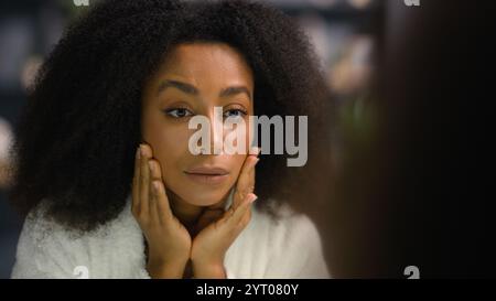 Gros plan miroir féminin reflet à la salle de bain de la maison de l'hôtel triste bouleversé mécontent femme afro-américaine mécontente vérifier mauvais teint problème rides Banque D'Images