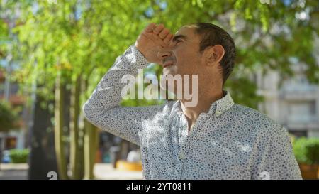 Homme hispanique d'âge moyen protégeant les yeux de la lumière du soleil dans un parc urbain, capturant un moment urbain en plein air avec un homme adulte mature au milieu de la verdure. Banque D'Images