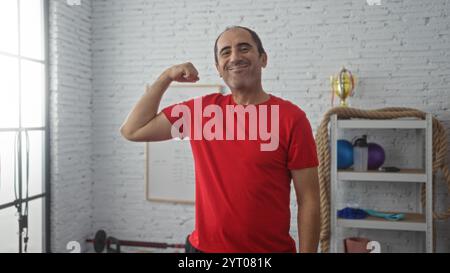 Homme hispanique d'âge moyen bras fléchissant dans la salle de gym avec des poids et trophée en arrière-plan Banque D'Images