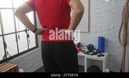 Homme hispanique debout avec les mains sur les hanches dans un intérieur de gymnastique, entouré d'appareils de fitness, sous une lumière intérieure brillante. Banque D'Images