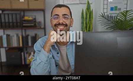 Jeune homme hispanique avec des lunettes et une moustache souriant dans un environnement de bureau avec un bureau soigneusement organisé et une étagère derrière lui Banque D'Images