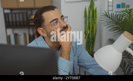 Jeune homme hispanique avec moustache et lunettes dans la salle de bureau pensant et se reposant la tête sur la main Banque D'Images