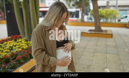 Femme enceinte aux cheveux blonds dans un parc urbain tenant doucement son ventre dans une lumière chaude reflétant la tranquillité de la grossesse au milieu de la vie urbaine Banque D'Images