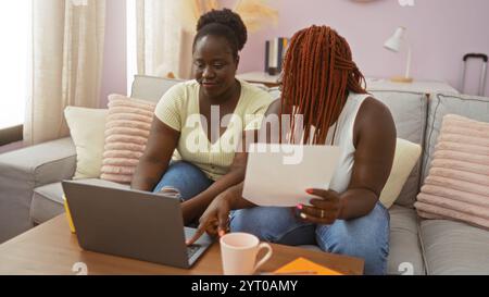 Deux femmes afro-américaines assises à l'intérieur dans un salon confortable travaillant ensemble sur un ordinateur portable et examinant un document. Banque D'Images