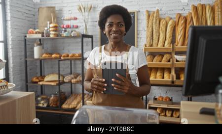 Femme tenant une tablette dans une boulangerie avec des étagères pleines de pain derrière elle, souriant et regardant la caméra, profitant de son travail dans un cadre intérieur lumineux. Banque D'Images