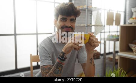 Bel homme hispanique à la barbe dégustant un café à l'intérieur dans un cadre de boulangerie, mettant en valeur des tatouages et souriant à la caméra Banque D'Images