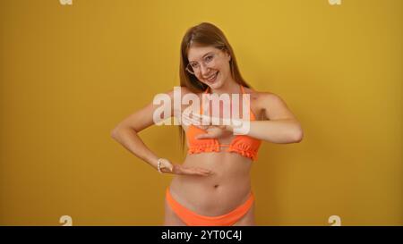 Jeune femme blonde attrayante dans un bikini orange posant avec confiance sur un fond jaune Banque D'Images