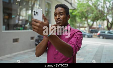 Jeune homme prenant selfie sur la rue urbaine portant chemise rose regardant réfléchi avec des voitures et des arbres en arrière-plan ambiance de ville vif expres énergique Banque D'Images
