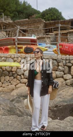 Jeune femme à cala deia, majorque, en plein air, portant des lunettes de soleil et une tenue d'été, debout près de kayaks, entourée de murs de pierre et de verdure Banque D'Images