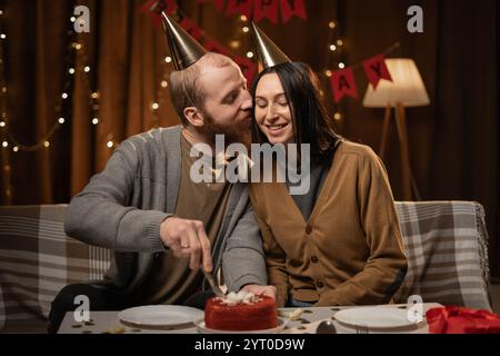 Couple dans l'amour portant des chapeaux de fête célébrant l'anniversaire à la maison mari coupant le gâteau sur l'anniversaire de la femme Banque D'Images