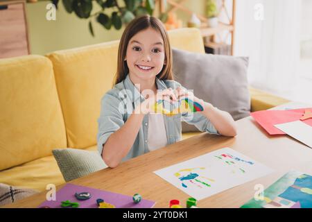 Jeune fille souriante tenant un cœur d'argile coloré à la maison dans un salon confortable, embrassant la créativité et le bonheur Banque D'Images